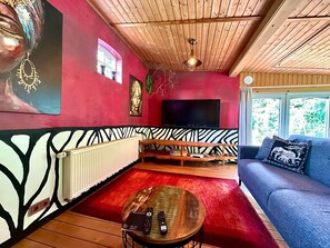 A room with a red wall, wooden ceiling, a large TV, a blue sofa, and a zebra-patterned bench. - Lodge of Africa (Hollern-Twielenfleth)