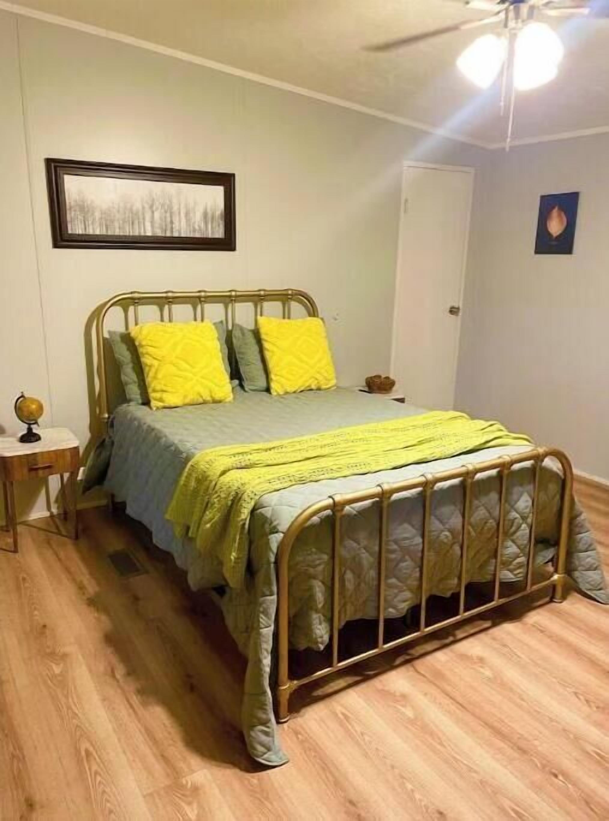 Een slaapkamer met een metalen bedframe, een houten nachtkastje met een wereldbol, en een ingelijst schilderij aan de muur.