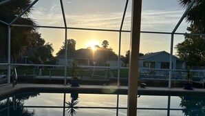 A pool area with a view of the sunset, surrounded by a glass enclosure and palm trees.