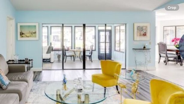 A spacious living room with a sofa, yellow chairs, and a glass coffee table. There is a dining area with a table and chairs visible through large glass doors.