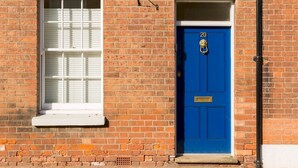 A brick building with a blue door marked with the number 20, a window with white shutters, and a metal pipe running along the wall.