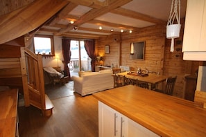 A wooden cabin interior with a dining area, a sofa, and a staircase leading to a balcony.