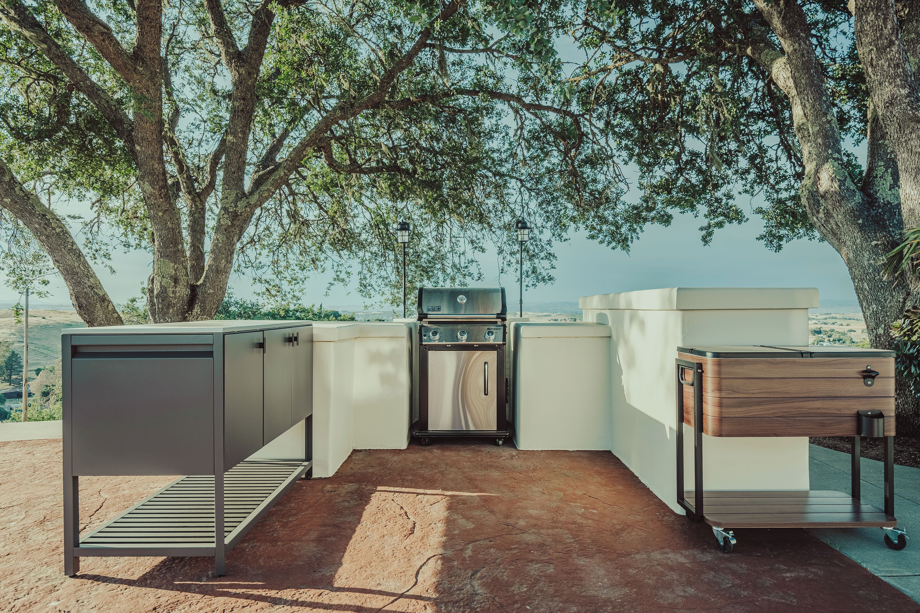 A compact outdoor kitchen setup with a built-in oven, a sink, and a storage cabinet, surrounded by trees and a scenic view.