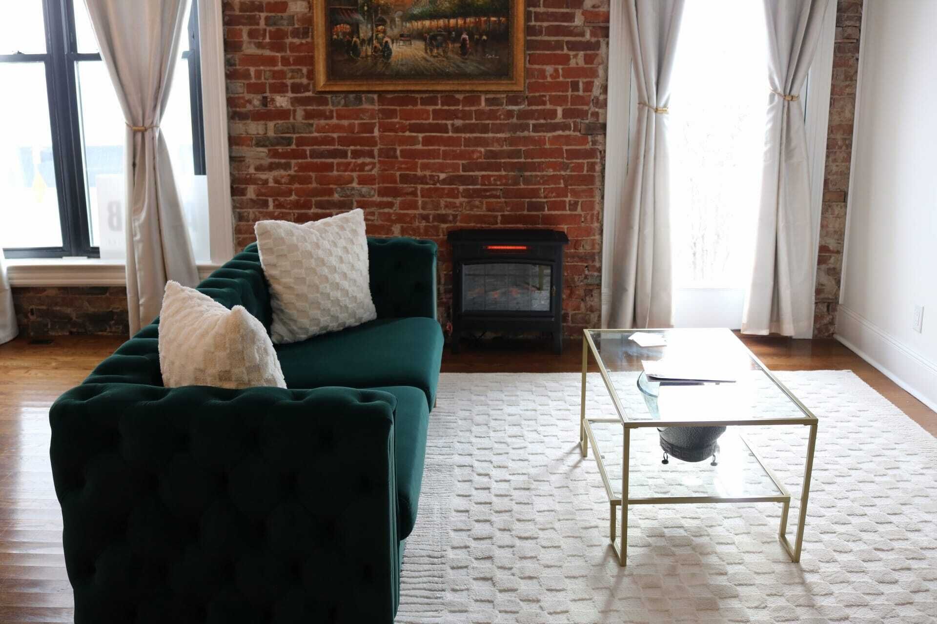 Una sala de estar con un sofá verde, cojines blancos, una mesa de centro de vidrio y una chimenea de ladrillo.
