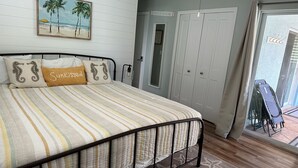 Un dormitorio con cama, un cuadro de playa, una puerta con la etiqueta 'BEACH HOUSE' y vista al exterior a través de una puerta de cristal.