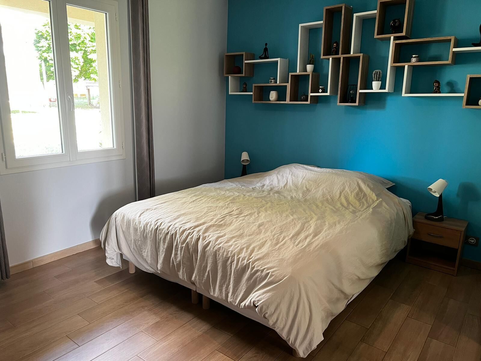 Un dormitorio con cama, mesitas de noche de madera, una pared azul como detalle y una ventana con cortinas.