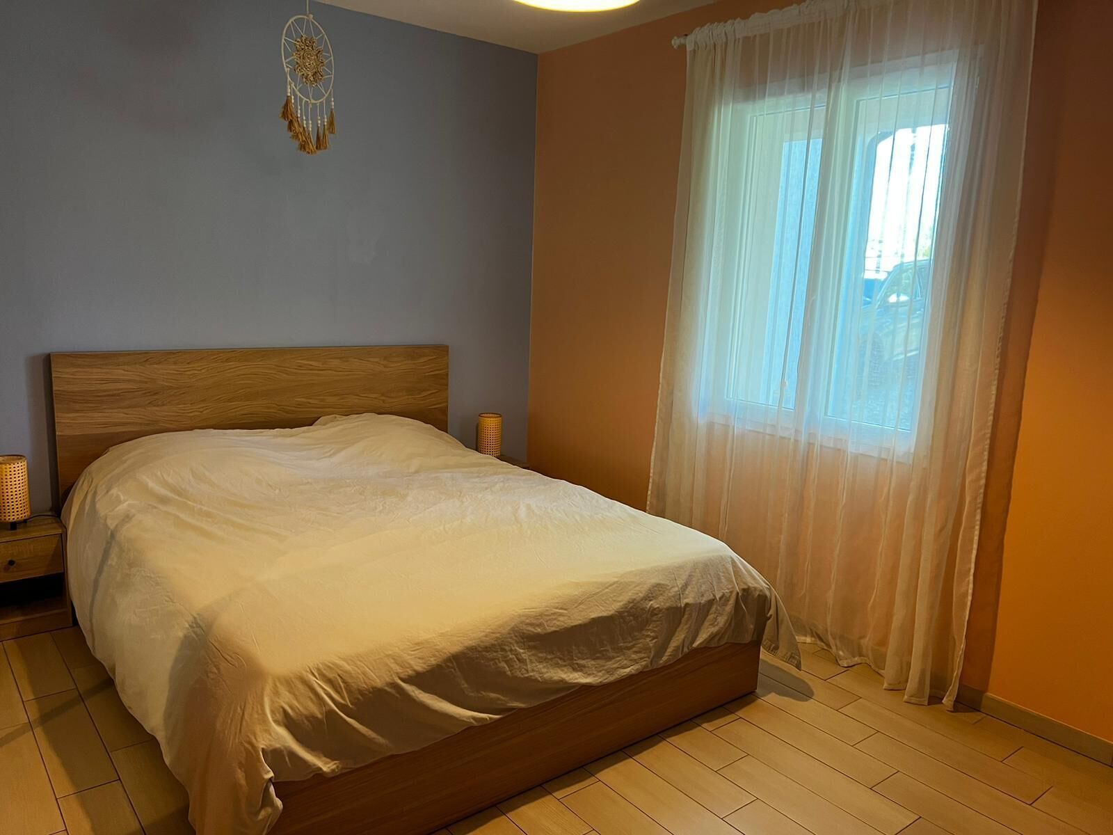 A bedroom with a wooden bed, a bedside lamp, a dreamcatcher on the wall, and sheer curtains over a window.