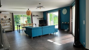 Un salon spacieux avec un canapé bleu, un coin repas comprenant une table et des chaises, un téléviseur et une grande fenêtre avec des rideaux.
