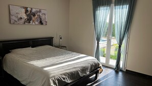 Une chambre avec un grand lit, une table de chevet, une lampe fixée au mur et une vue sur une piscine à travers des rideaux transparents.