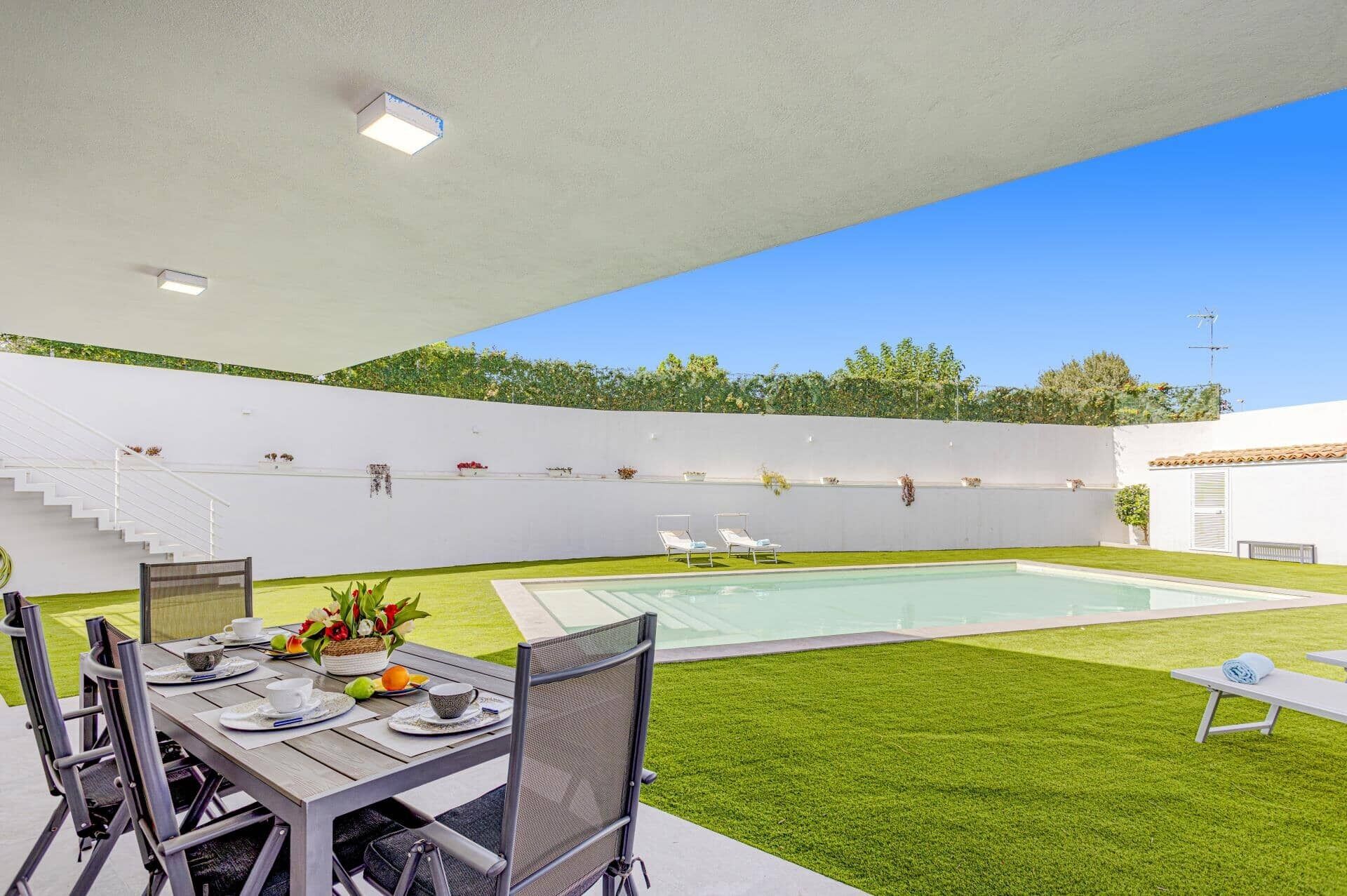 Un área de comedor exterior moderna con piscina, paredes blancas y una escalera que conduce a un nivel superior.