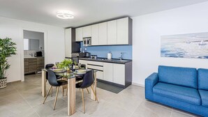 Een moderne keuken met witte kasten, een ingebouwde magnetron en een blauwe spatwand. In de woonkamer staat een eettafel met stoelen en een blauwe bank.