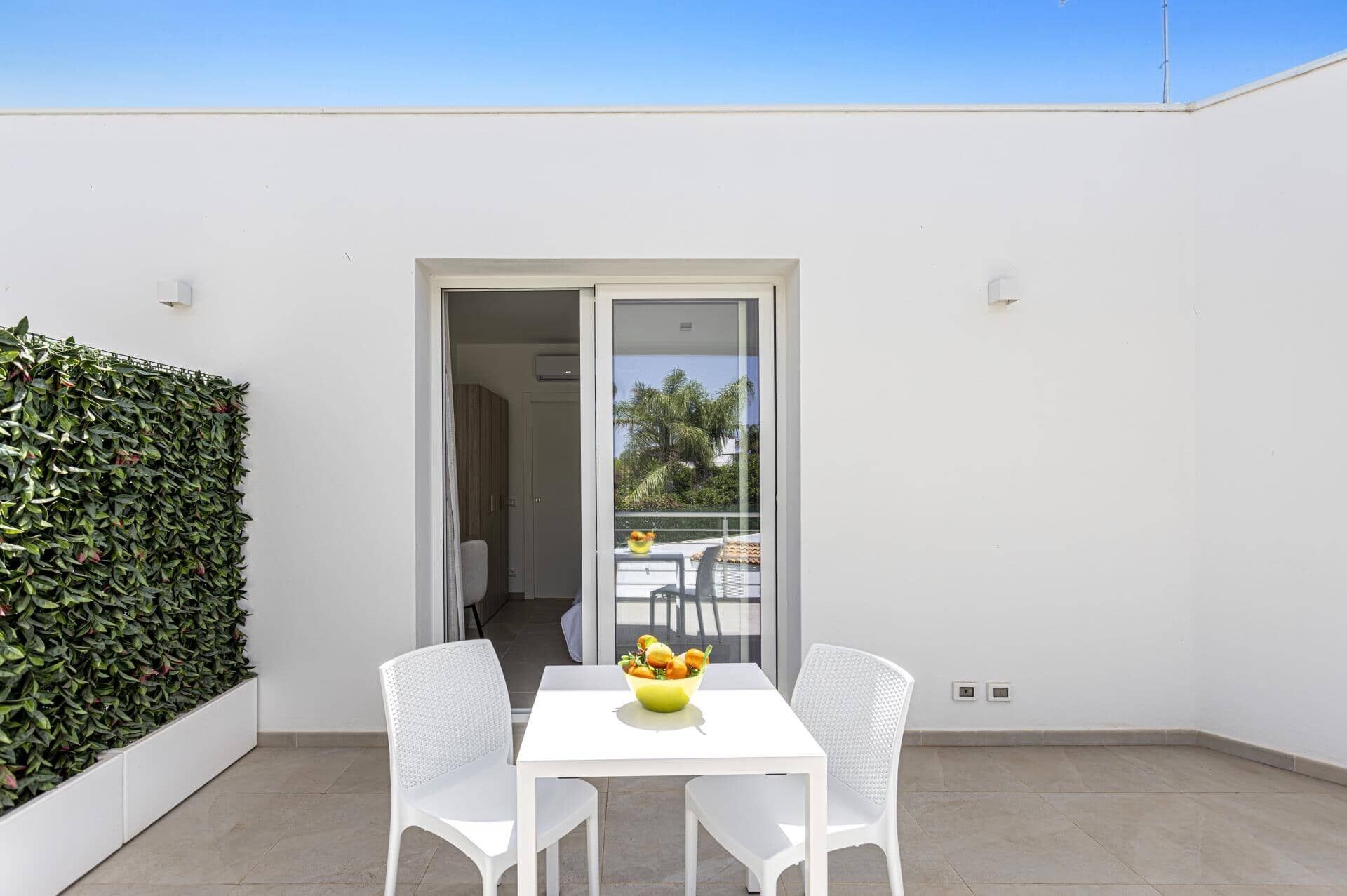 A balcony with a white table and two white chairs, a glass door leading to an indoor area, and a green hedge on the left.