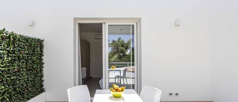 A balcony with a white table and two white chairs, a glass door leading to an indoor area, and a green hedge on the left.