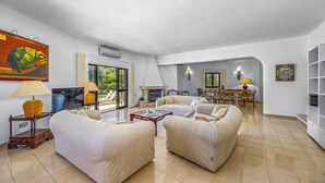 A spacious living room with a fireplace, a dining area, and a large window offering a view of the outdoors. - Villa Casa Jasmim by Villa Plus (Quarteira)