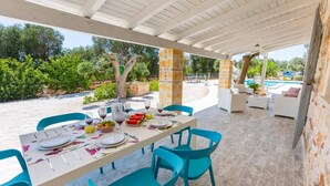 An outdoor dining area with a table set for a meal, surrounded by chairs, and a pool in the background. - Villa Trullo Famigiu by Villa Plus (Grottaglie)