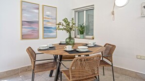 A dining area with a round wooden table, four wicker chairs, a vase with plants, and two framed artworks on the wall.