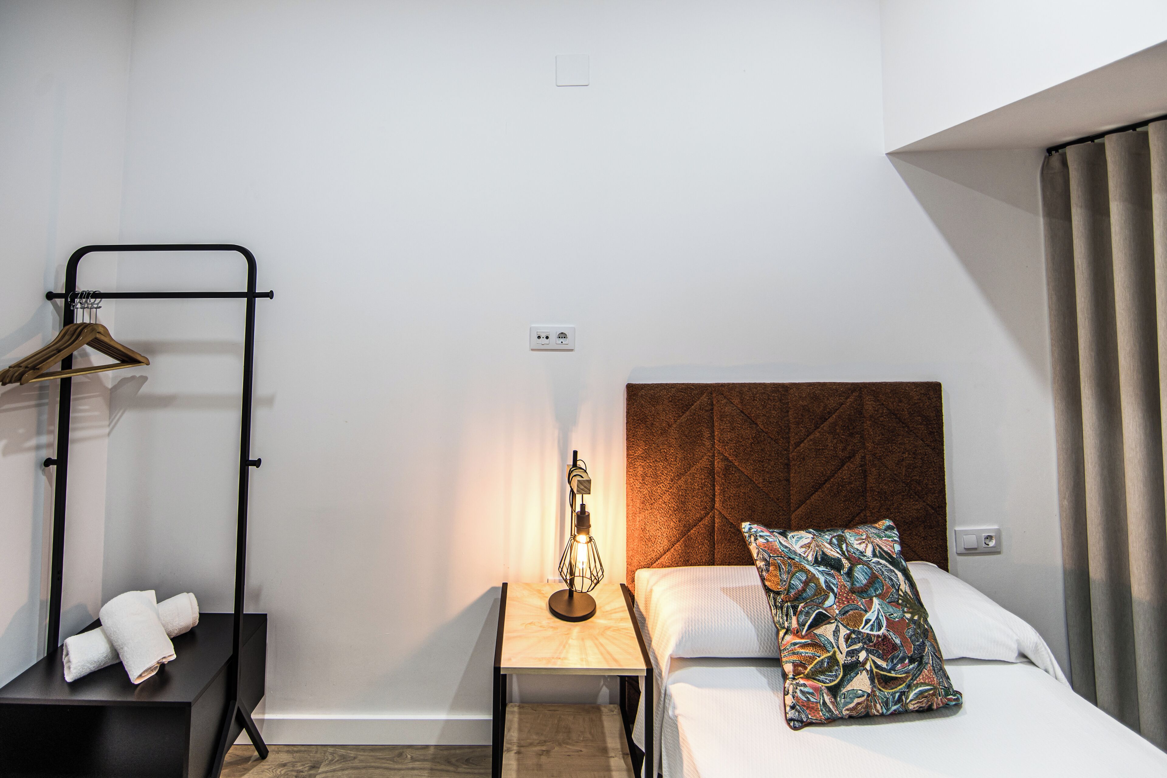 A hotel room with a bed, a bedside table with a lamp, a towel rack, and a hanging coat rack.