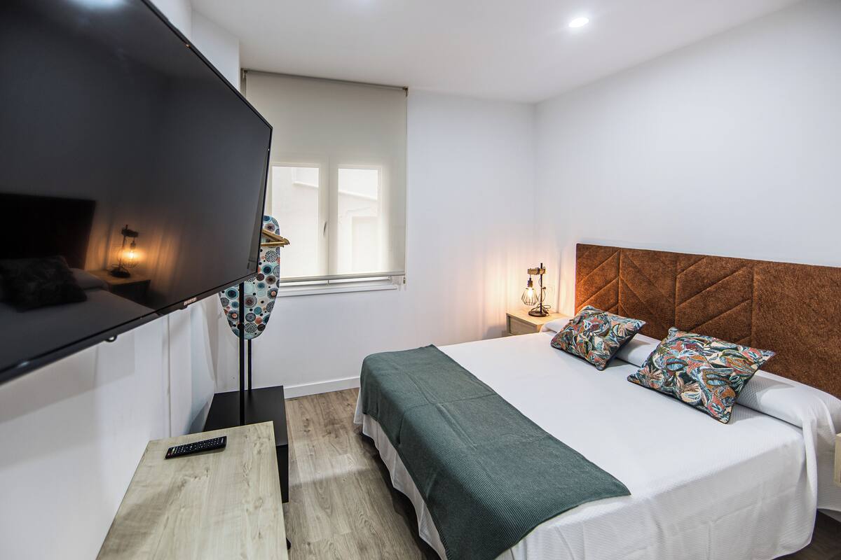 Une chambre à coucher avec un lit, une télévision, une table de chevet, une lampe et une fenêtre.