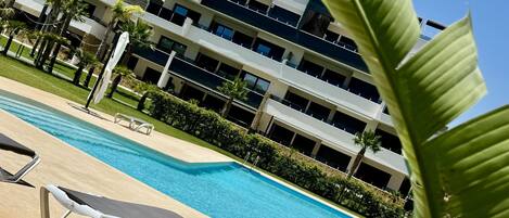 Una piscina con sillas de descanso, rodeada de vegetación y un edificio moderno con varios balcones.