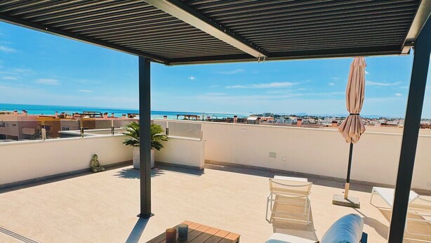A rooftop terrace with a blue sofa, a wooden table, and a view of the beach.