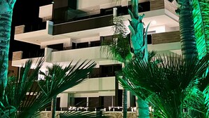 A modern multi-story building with balconies, surrounded by palm trees and illuminated by green lights.