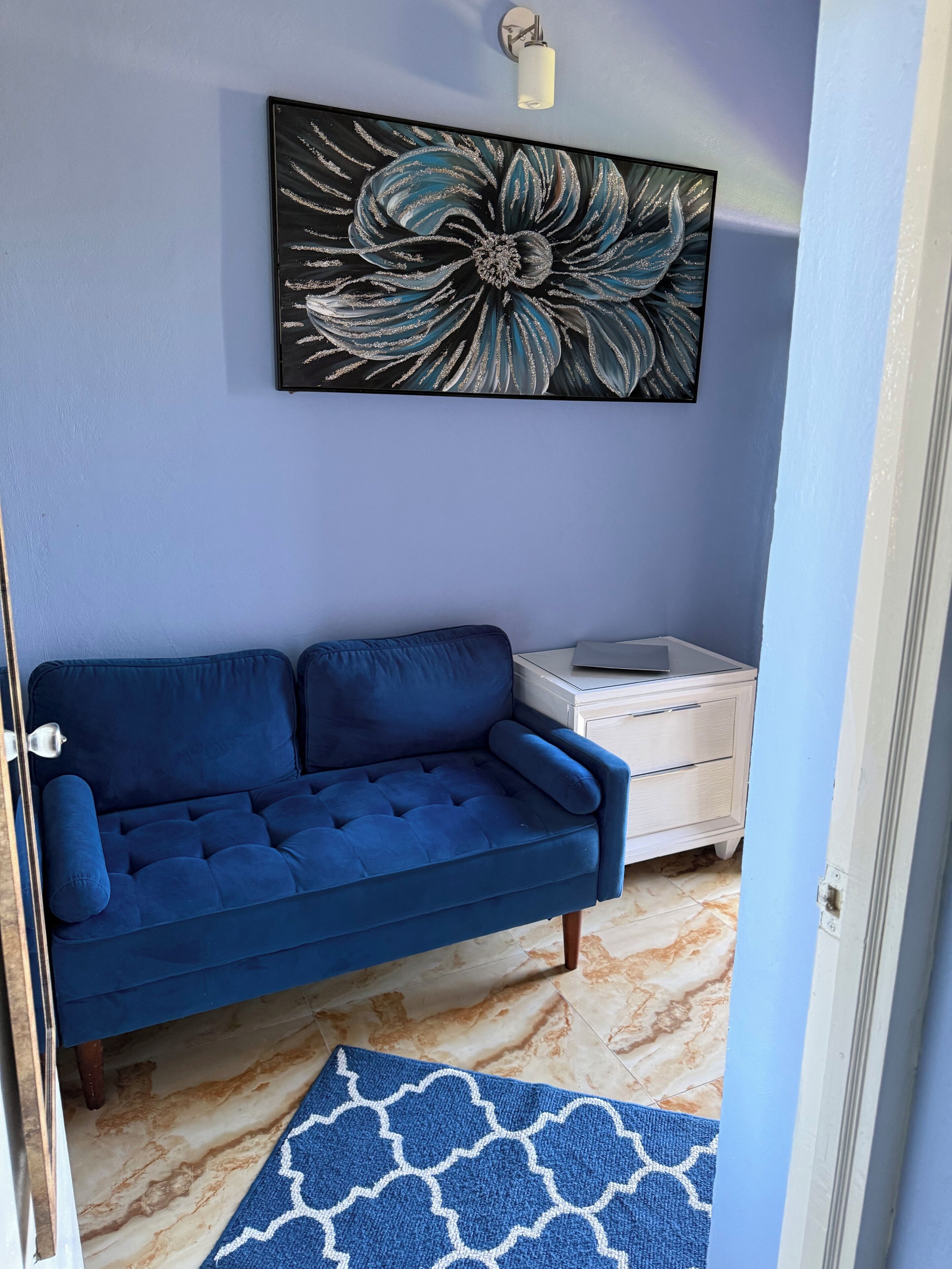 Una habitación con un sofá azul, un adorno en la pared, una mesita de noche pequeña y una alfombra estampada.