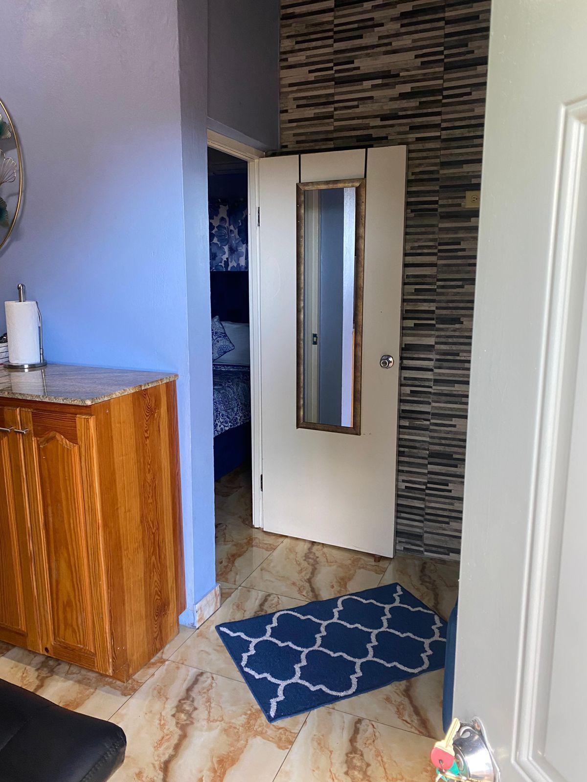 A room with a wooden wardrobe, a white door with a mirror, a blue patterned rug, and a tiled floor.