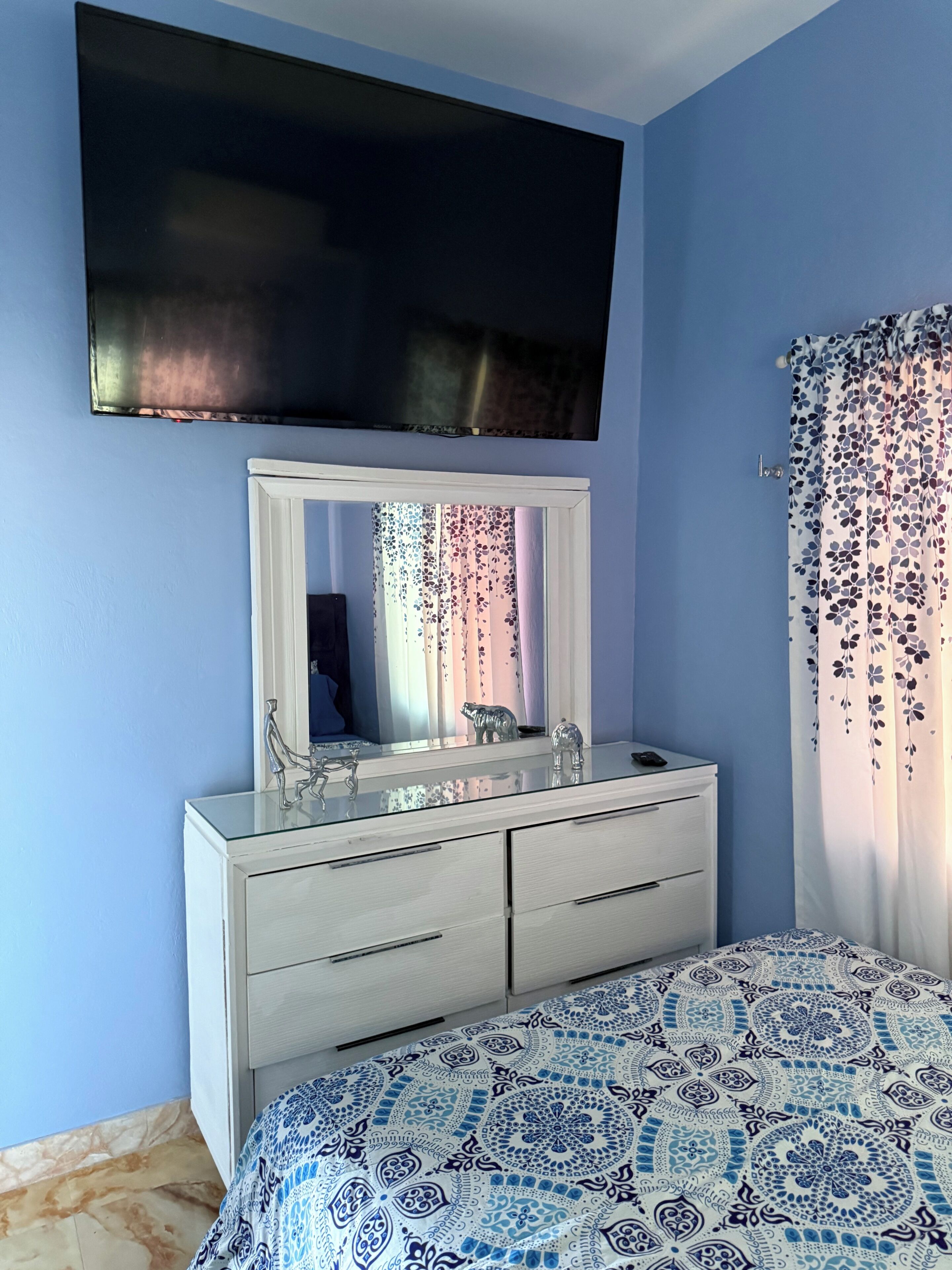 A bedroom with a bed, a dresser, a mirror, a flat-screen TV, and a window with a floral curtain.