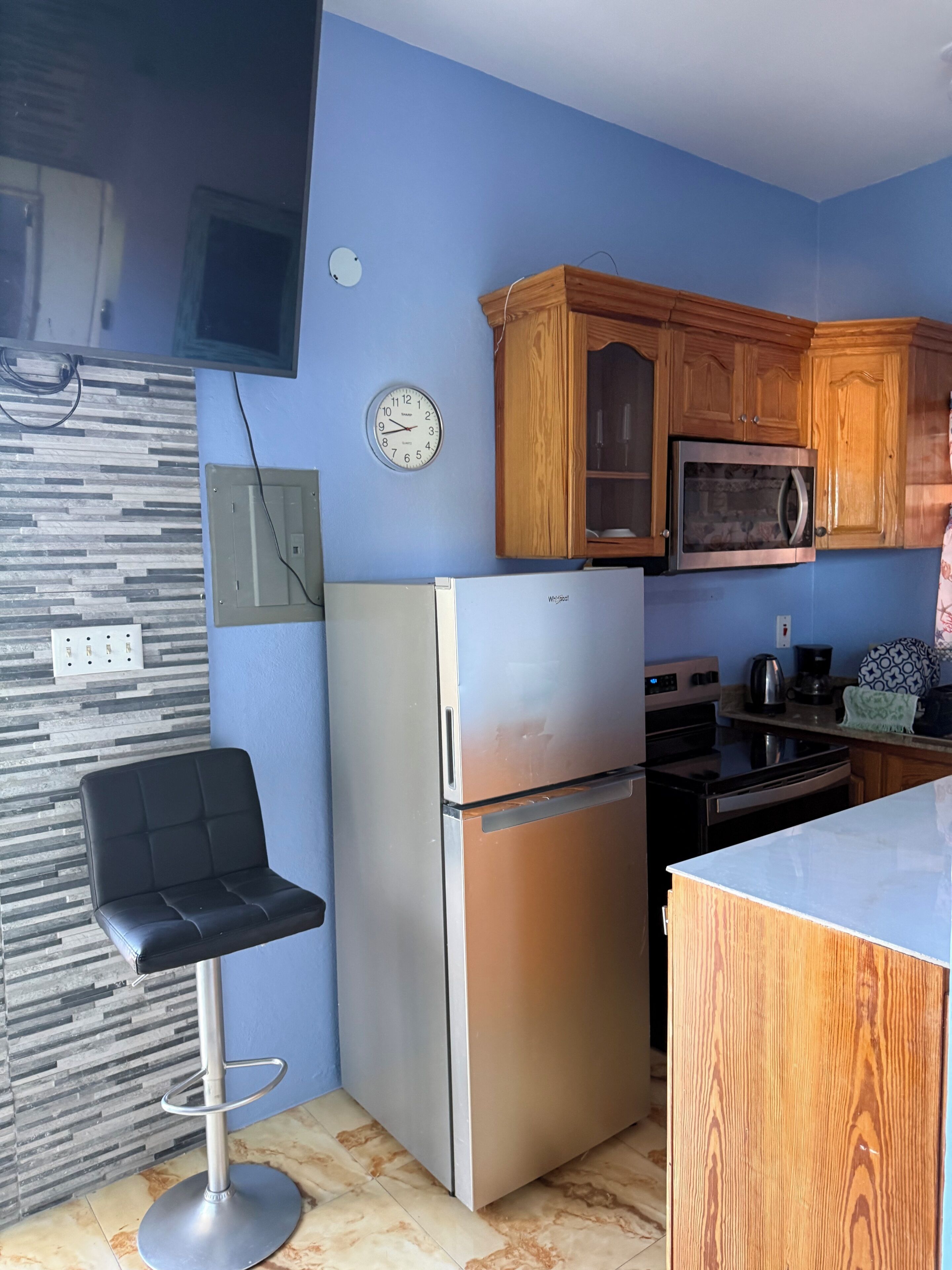 Una cocina con refrigerador, microondas, armarios de madera, un reloj y un taburete de bar.