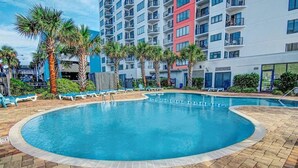 A swimming pool with lounge chairs, palm trees, and a multi-story building with balconies in the background.