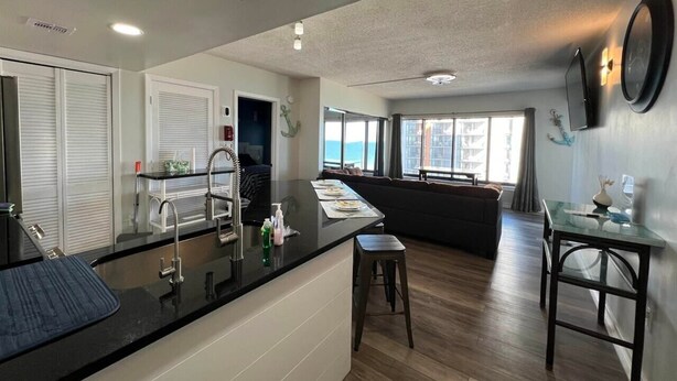 A modern kitchen with a black countertop, stainless steel sink, and a dining area with a glass table and bar stools.