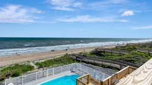 Une vue sur la plage avec une terrasse en bois, une piscine et une zone de plage où se trouvent des personnes et des parasols.