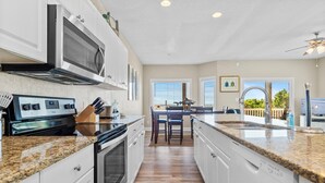 Una cocina moderna con armarios blancos, encimeras de granito, electrodomésticos de acero inoxidable y vistas al exterior.