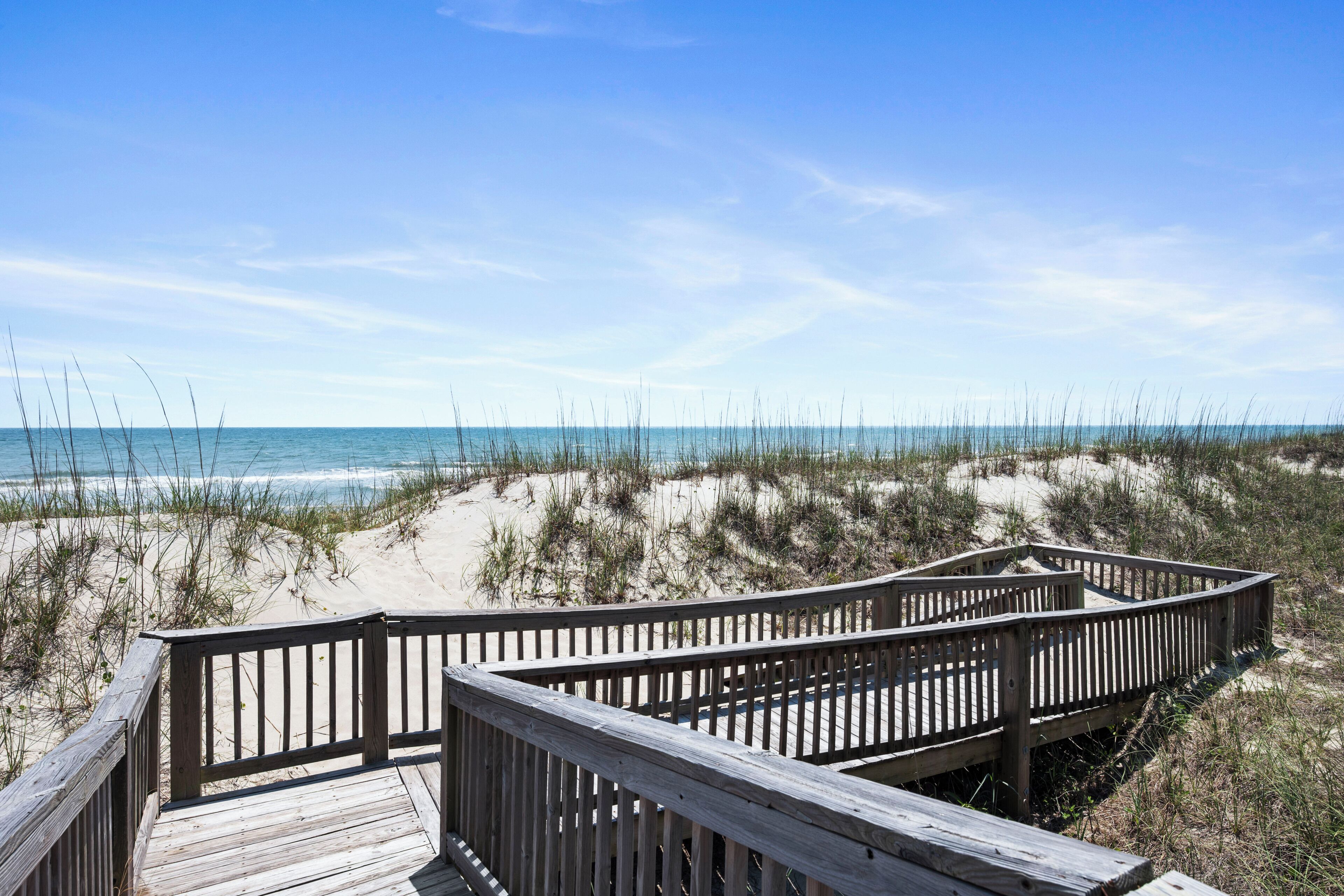 A wooden boardwalk leading to a beach with sand dunes and sparse vegetation.