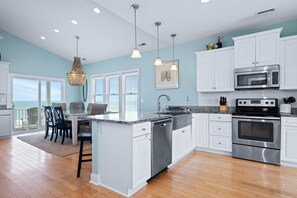 A kitchen with white cabinets, stainless steel appliances, a dining area with chairs, and a view through the window. - Sea La Vie- Oceanfront-Secluded-beautiful! Private pool and hot tub! (Emerald Isle)