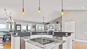 A modern kitchen with a central island, white cabinetry, and a view of the ocean through large windows. - Oceanfront 8 Bedroom/6.5 Bath Home: Elevator, Gourmet Kitchen, Pool, Hot Tub, St (Emerald Isle)