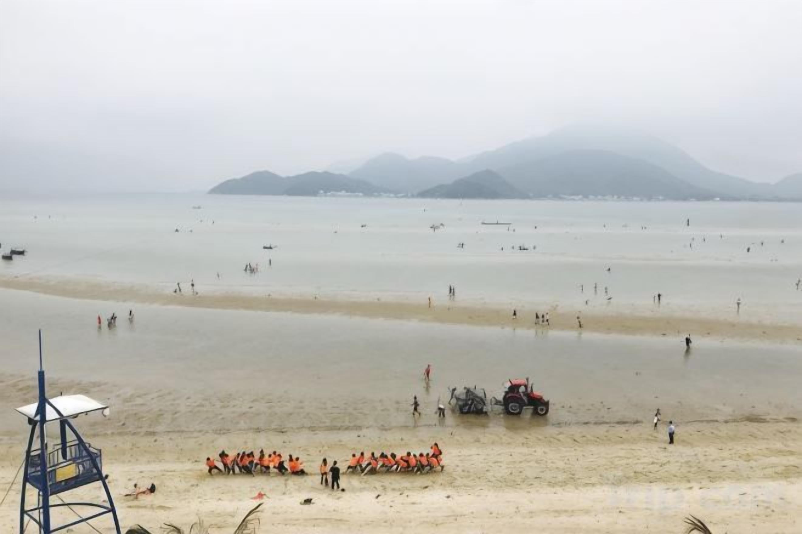 Una playa con una torre de salvavidas, gente con chalecos anaranjados y un tractor sobre la arena.