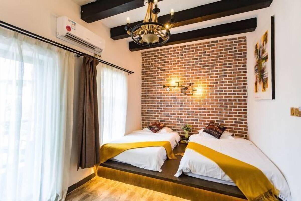 Habitación de hotel con dos camas, pared de ladrillo, candelabro y ventana con cortinas.