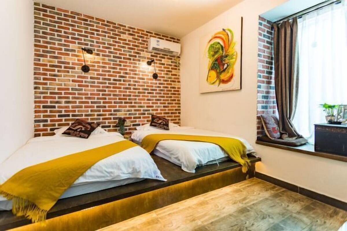 Una habitación con dos camas, una pared de ladrillo, un cuadro abstracto colorido y una ventana con cortinas.