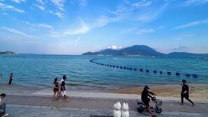 Une promenade en bord de mer où l’on voit des gens se promener et faire de la trottinette, un ciel dégagé et une montagne lointaine.