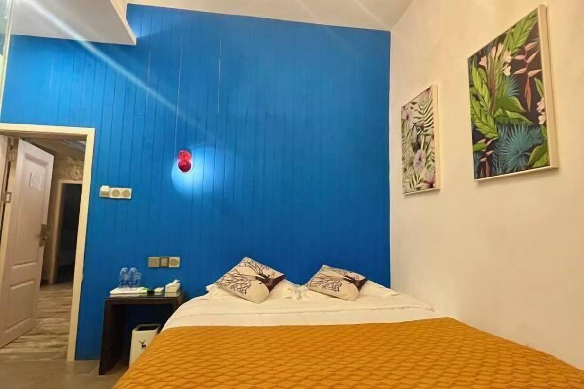 Una habitación de hotel con pared azul, una cama con ropa de cama blanca y almohadas marrones, una mesita de noche con una lámpara y un cuadro de hojas tropicales.