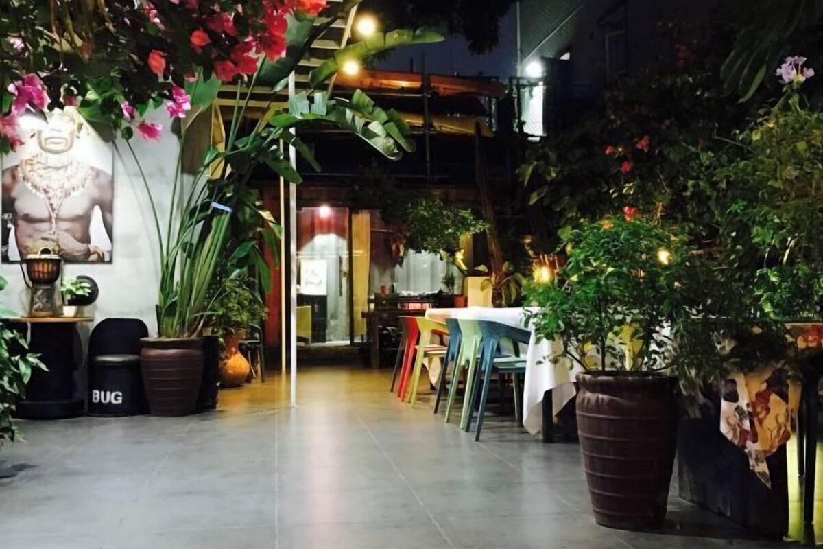 An indoor dining area with tables and chairs, potted plants, and a wall-mounted light fixture.