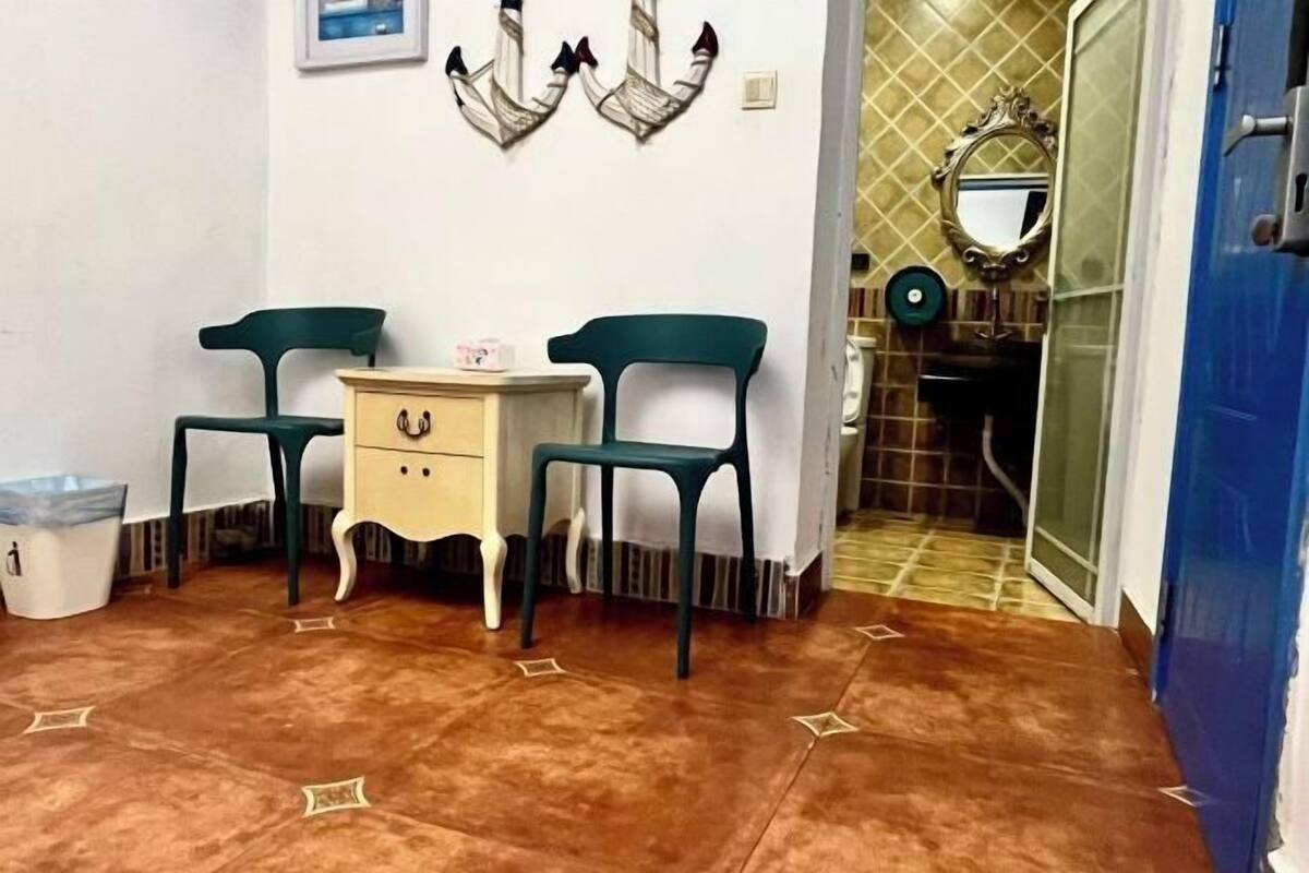 Una habitación pequeña con dos sillas, una mesita con cajones, un cubo de basura y un espejo en la pared.