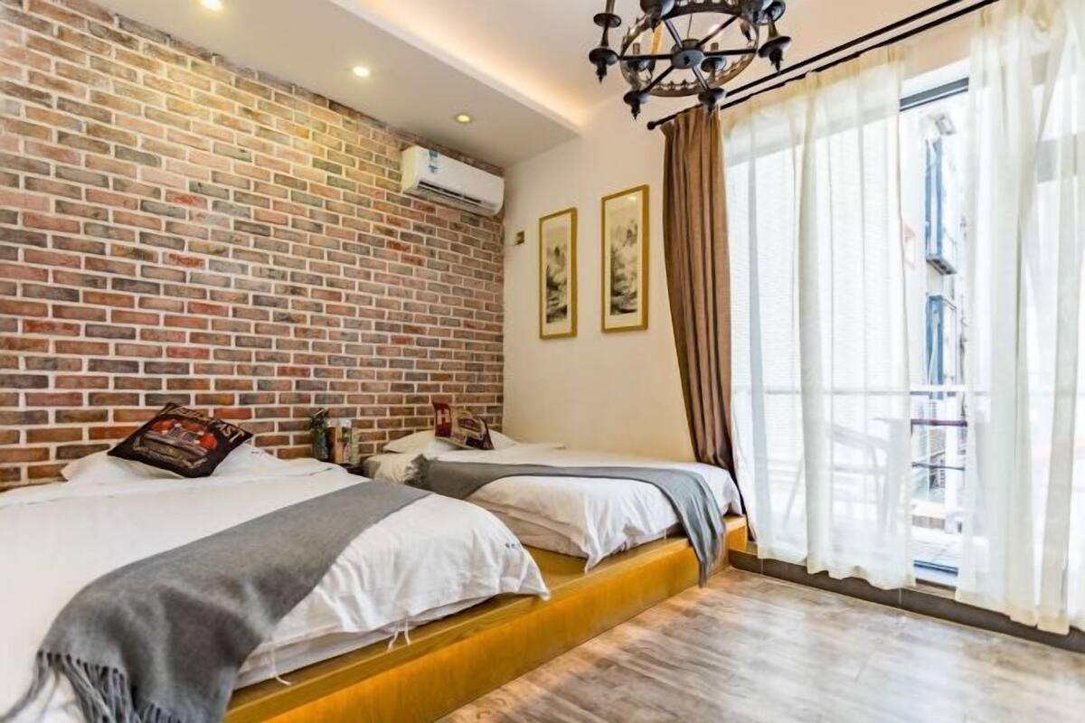 Una habitación con dos camas, una pared de ladrillo y un ventanal con cortinas.