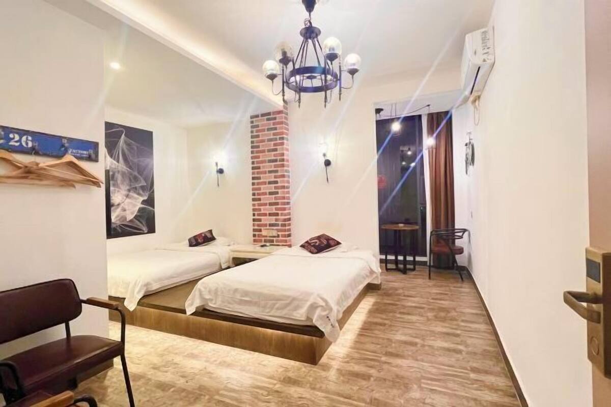 Habitación de hotel con dos camas, un banco de madera, una araña y una pared de ladrillo decorativa, además de vistas al exterior.