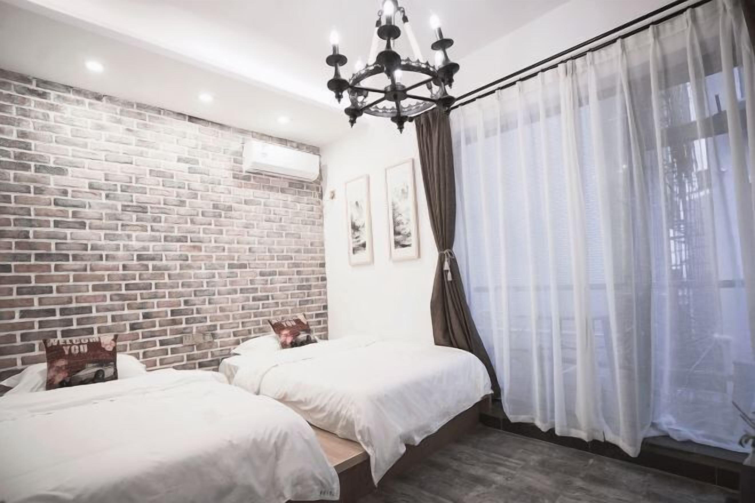 Una habitación con dos camas, una pared de ladrillo, una araña y una ventana con cortinas traslúcidas.