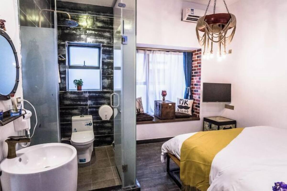 Una habitación de hotel moderna con una cama grande, un baño con cabina de ducha de vidrio, un ventanal con vistas y una lámpara de araña original.
