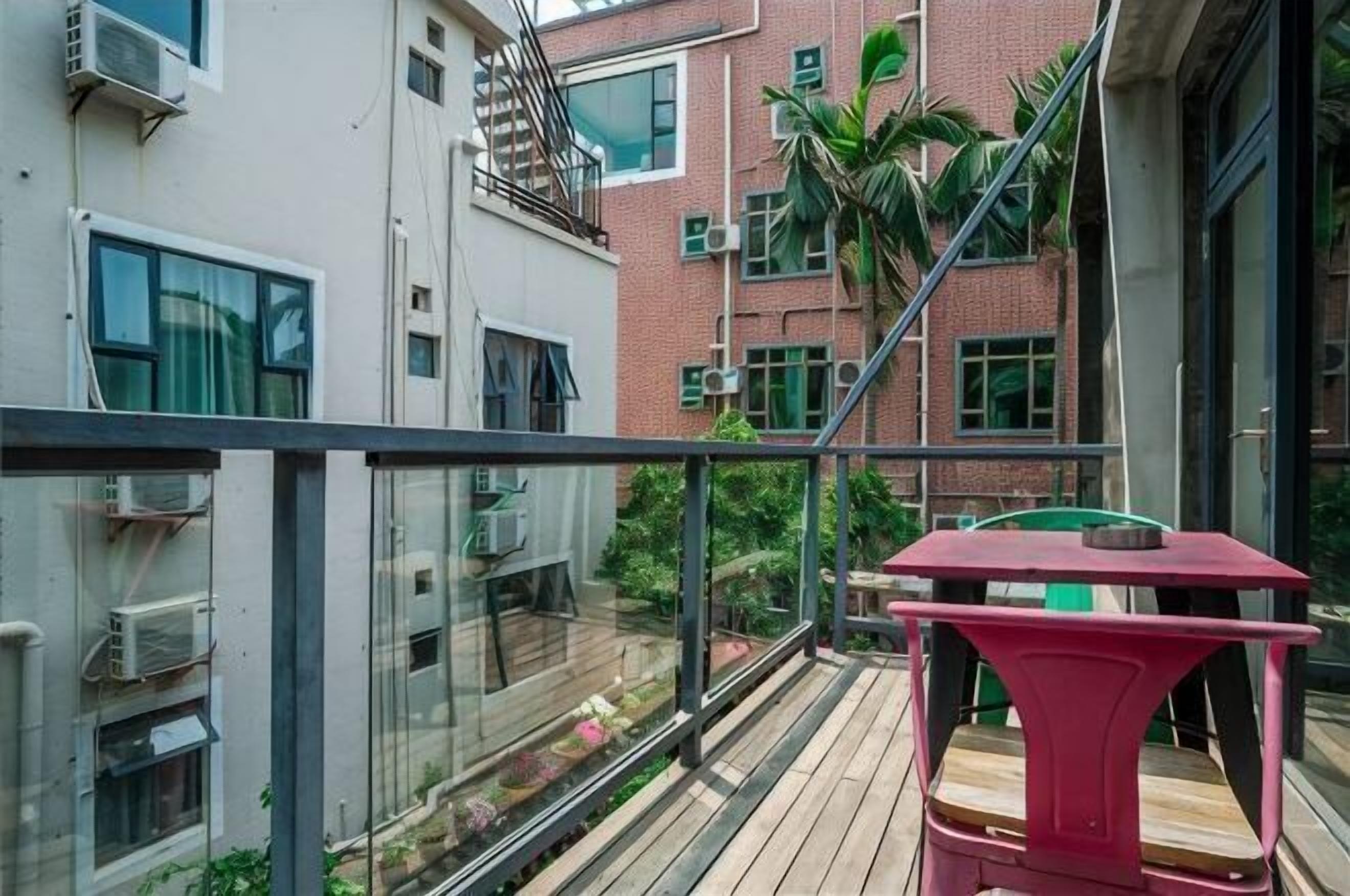 A balcony with a red table, glass railings, and a view of a brick building with multiple windows and air conditioning units.
