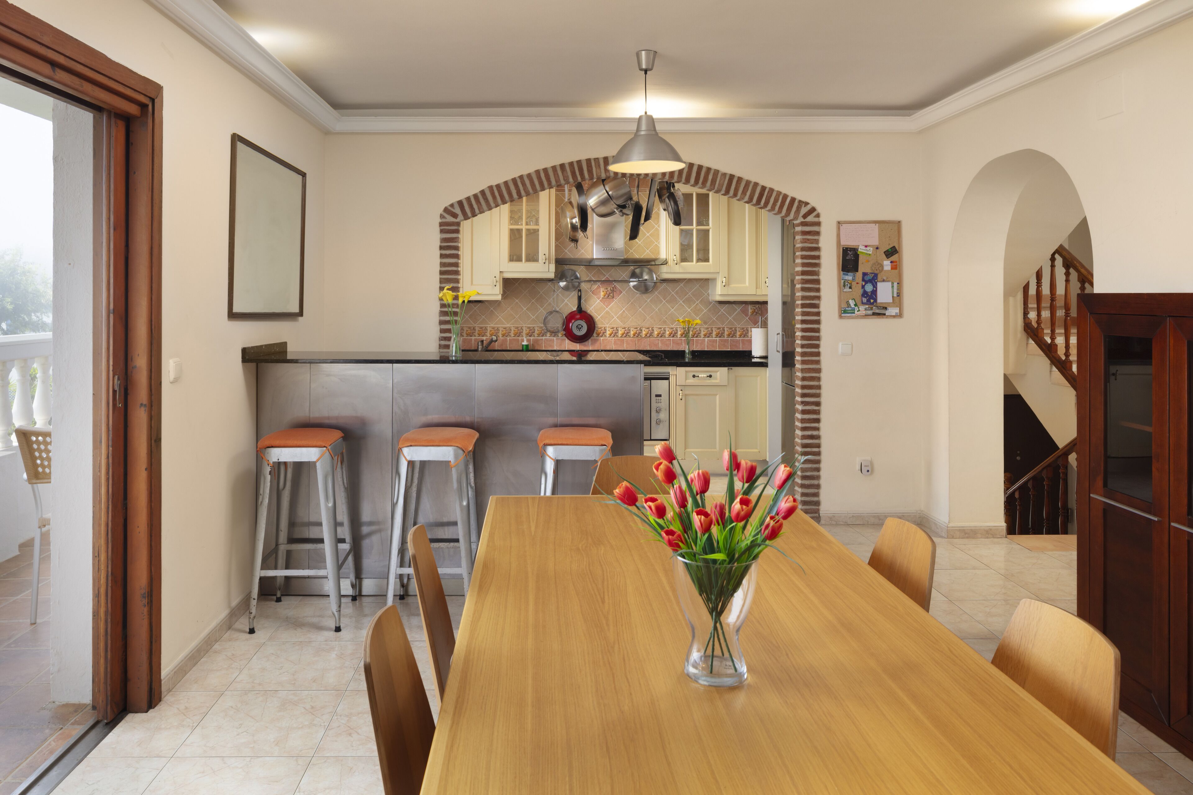 Een eethoek met een houten tafel, stoelen en een vaas tulpen. Er is een keuken met een bakstenen boog, kasten en een hangende pannendroger.