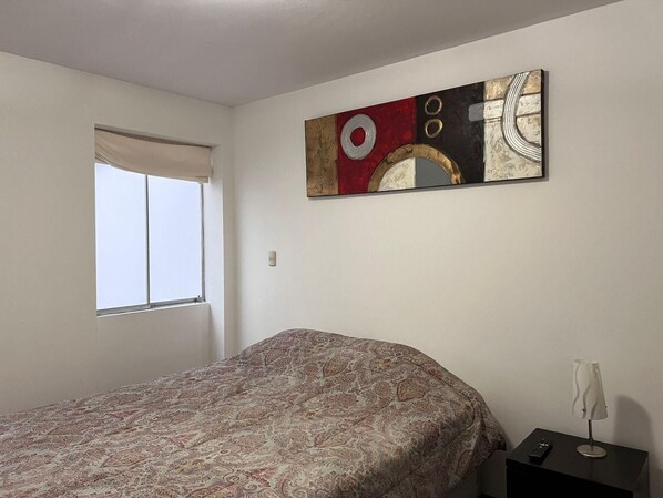 A bedroom with a bed, a window, a wall-mounted abstract painting, and a bedside table with a lamp. - 3bed Apt in Miraflores - Free Wifi, Free Parking (Lima)